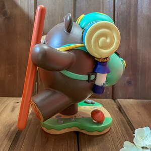May include: A brown bear figurine with a green and yellow backpack, holding a red stick. The bear is standing on a green base with a mushroom and rocks.