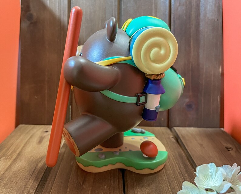 May include: A brown bear figurine with a green and yellow backpack, holding a red stick. The bear is standing on a green base with a mushroom and rocks.