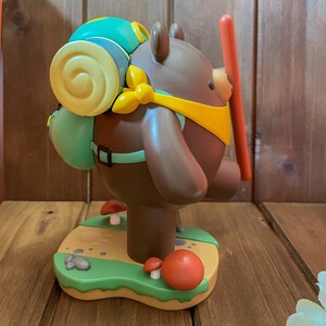 May include: A brown bear figurine with a yellow bandana and a green backpack. The bear is standing on a green base with brown rocks and red mushrooms.