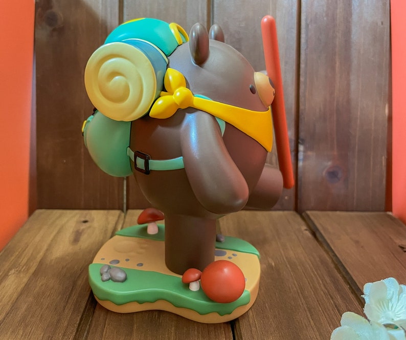 May include: A brown bear figurine with a yellow bandana and a green backpack. The bear is standing on a green base with brown rocks and red mushrooms.