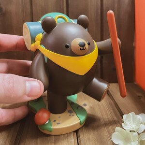 May include: A brown bear figurine with a yellow scarf and a green backpack, holding a stick. The bear is standing on a brown base with green and brown details.