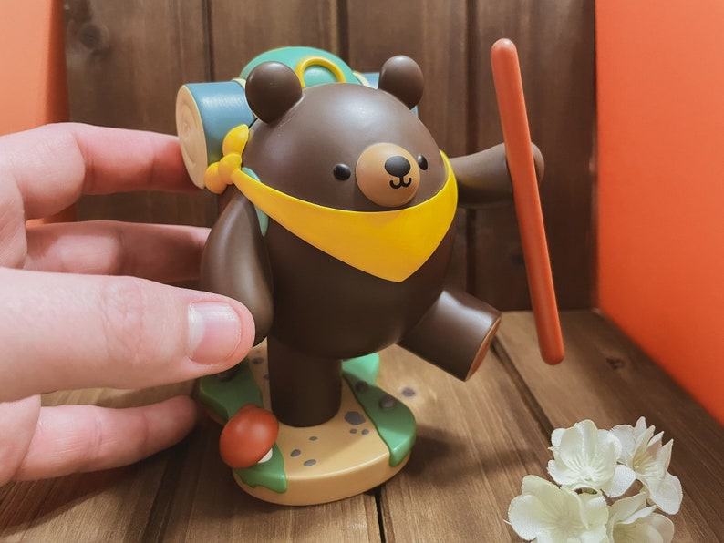 May include: A brown bear figurine with a yellow scarf and a green backpack, holding a stick. The bear is standing on a brown base with green and brown details.