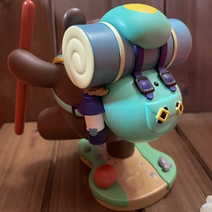 May include: A brown bear figurine with a blue and yellow backpack, a red stick, and a blue water bottle. The bear is standing on a green and brown base.