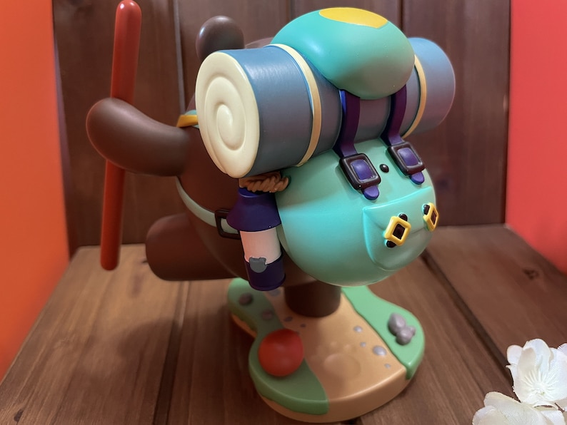 May include: A brown bear figurine with a blue and yellow backpack, a red stick, and a blue water bottle. The bear is standing on a green and brown base.