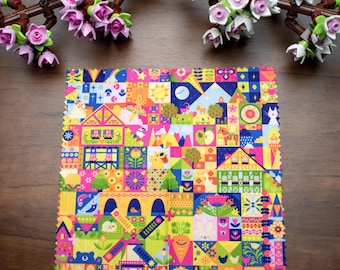 May include: A colorful square fabric with a patchwork design featuring houses, animals, and floral elements. The fabric has a vibrant mix of pink, yellow, blue, and green hues. Decorative branches with pink and white flowers are in the background.