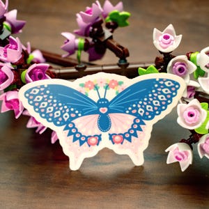 May include: A sticker featuring a blue and pink butterfly with floral accents. The butterfly has detailed wing patterns and is surrounded by artificial pink flowers and branches.