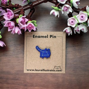 May include: A blue enamel pin in the shape of a cat with a constellation design. The pin is on a brown card that reads "Enamel Pin" and "www.laurailustrates.com". The background includes a wooden surface and a branch with pink and white flowers.