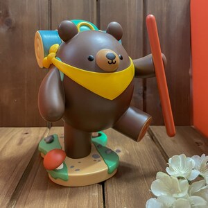 May include: A brown bear figurine with a yellow scarf and a green backpack, holding a red stick. The bear is standing on a green and brown base with a mushroom.