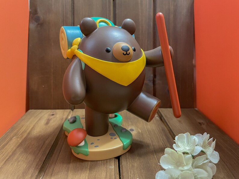 May include: A brown bear figurine with a yellow scarf and a green backpack, holding a red stick. The bear is standing on a green and brown base with a mushroom.