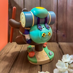 May include: A brown and green figurine of a bear wearing a blue backpack with a yellow patch. The bear is holding a stick in its right paw and is standing on a green base with a brown path.
