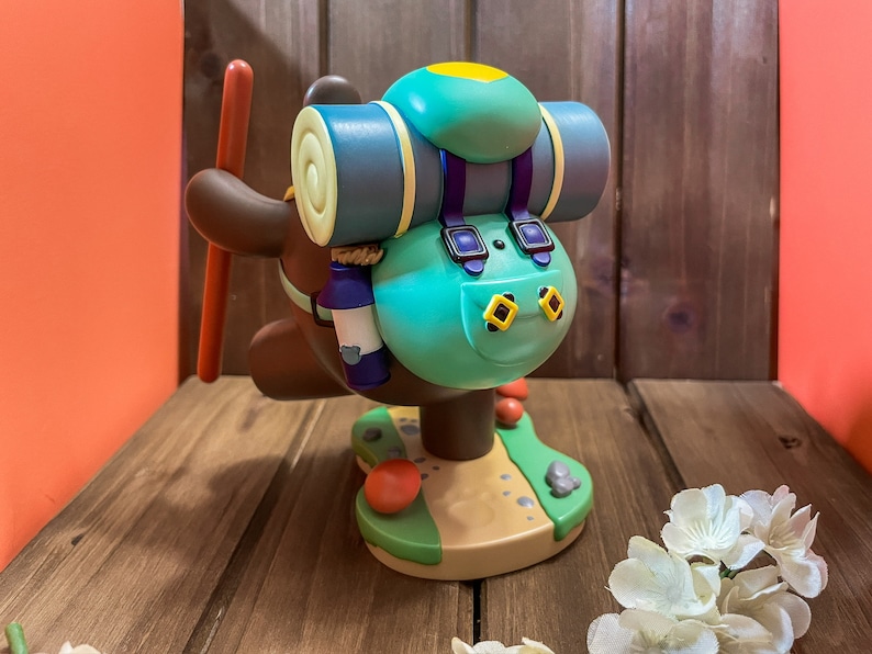 May include: A brown and green figurine of a bear wearing a blue backpack with a yellow patch. The bear is holding a stick in its right paw and is standing on a green base with a brown path.