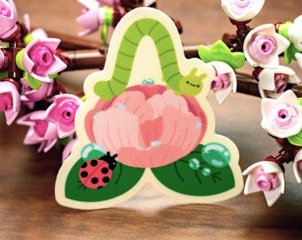 May include: A whimsical sticker featuring a pink peony, a green caterpillar, and a red ladybug. The sticker is surrounded by artificial pink flowers with white petals and brown stems, creating a decorative floral arrangement.
