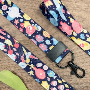 May include: Navy blue lanyards with a repeating pattern of colorful sea creatures, including fish, jellyfish, and starfish. Each lanyard has a black clasp and a black rectangular connector. The lanyards are on a wooden surface.