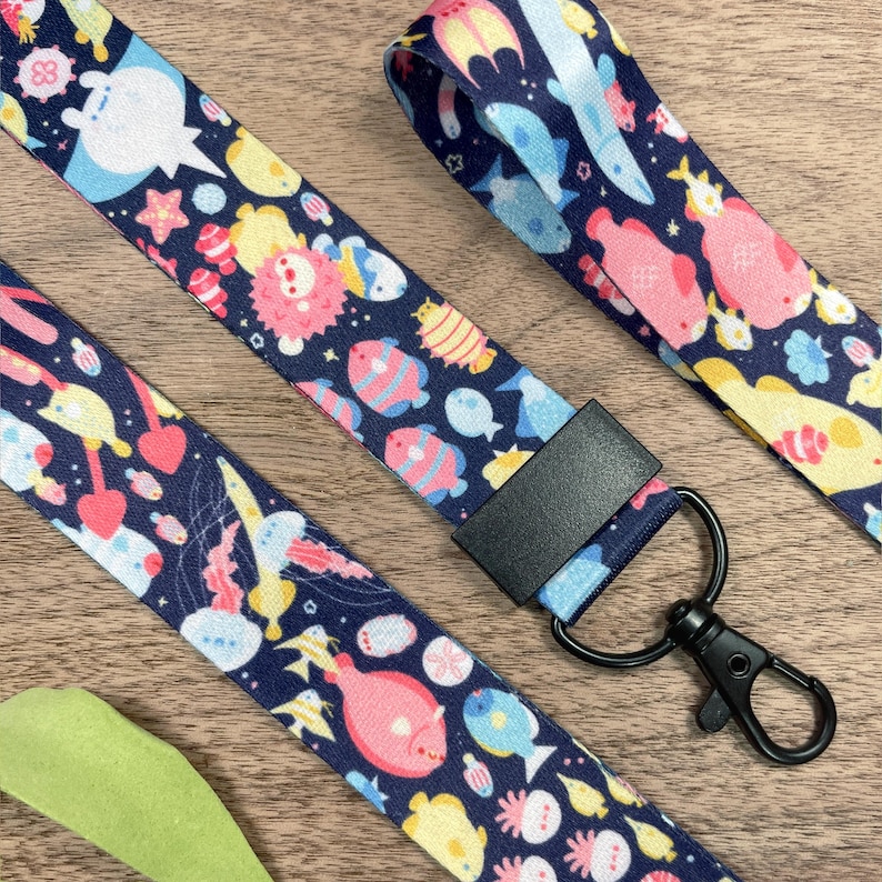 May include: Navy blue lanyards with a repeating pattern of colorful sea creatures, including fish, jellyfish, and starfish. Each lanyard has a black clasp and a black rectangular connector. The lanyards are on a wooden surface.