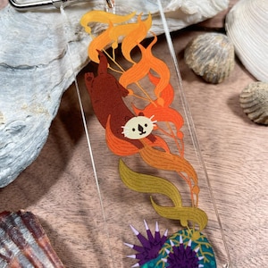 May include: A clear acrylic keychain featuring a cartoon otter swimming among orange and yellow seaweed. The otter is brown with a white face and black details. The keychain has a silver clasp.