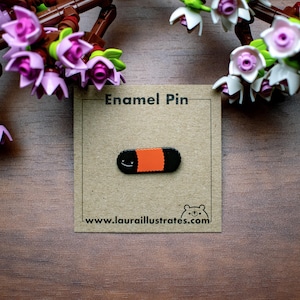 May include: An enamel pin featuring a sushi roll design with a smiling face. The pin has a black base with an orange center and is displayed on a brown card that reads "Enamel Pin" and "www.laurailustrates.com". The background includes artificial flowers.
