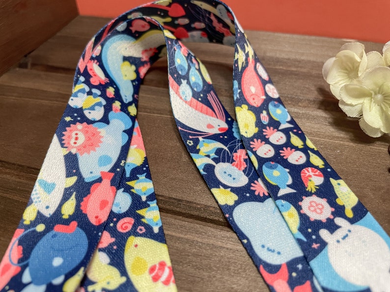May include: Navy blue lanyard with a repeating pattern of whimsical sea creatures in shades of pink, yellow, and blue. The lanyard is made of a smooth, durable material, perfect for holding keys or an ID badge.