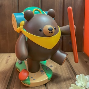 May include: A brown bear figurine with a yellow scarf and a green backpack, holding a red stick. The bear is standing on a green and brown base with a mushroom.