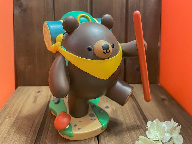 May include: A brown bear figurine with a yellow scarf and a green backpack, holding a red stick. The bear is standing on a green and brown base with a mushroom.