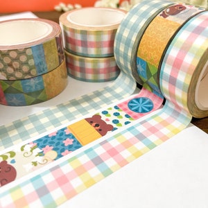 May include: A collection of decorative washi tape rolls in various patterns and colors. Designs include gingham, floral, and bear motifs. The tape is displayed on a white surface, ideal for crafts and scrapbooking.