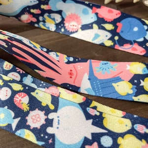 May include: Close-up of three patterned ribbons featuring an underwater theme. The ribbons have a dark blue background with colorful illustrations of various sea creatures, including fish, jellyfish, and whales. The ribbons are made of a smooth, shiny material.