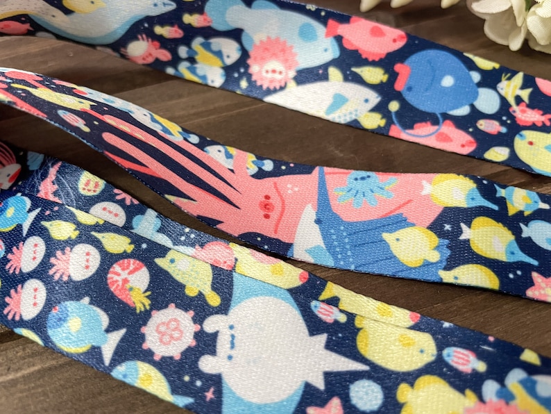 May include: Close-up of three patterned ribbons featuring an underwater theme. The ribbons have a dark blue background with colorful illustrations of various sea creatures, including fish, jellyfish, and whales. The ribbons are made of a smooth, shiny material.