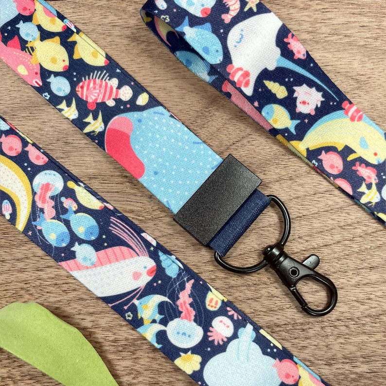 May include: Navy blue lanyards with a colorful sea life pattern. The lanyards feature various marine creatures like fish, jellyfish, and stingrays. Each lanyard has a black metal clasp and a dark blue fabric piece.
