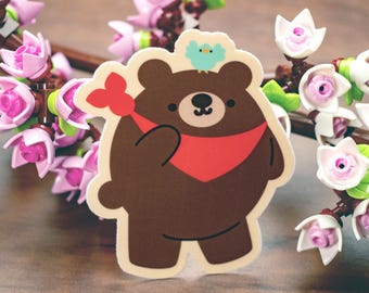 May include: A brown bear sticker with a red scarf and a small blue bird on its head. The bear is holding a red flower. The sticker has a white border and is set against a background of pink flowers.
