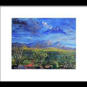May include: Framed landscape painting depicting a desert scene under a blue sky with clouds. The artwork features mountains in the distance, green vegetation, and a variety of cacti. The painting is framed in a black frame.