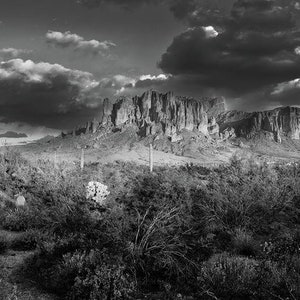May include: A black and white photograph of a mountain range with a dramatic sky. The mountain range is in the distance, and the sky is filled with dark clouds. The foreground is a field of brush.