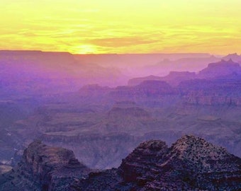 May include: A panoramic view of a canyon at sunset, with the sun setting behind the distant mountains. The canyon is filled with layers of rock formations, and the sky is a vibrant mix of yellow, orange, and purple.