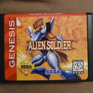 May include: A Sega Genesis game cartridge for Alien Soldier. The cartridge is red and black with a yellow and orange background image of a robot with a bird-like head. The game is rated "KA" for Kids to Adults by the ESRB.