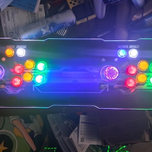 May include: A rectangular arcade game controller featuring a black surface with illuminated buttons in red, yellow, green, blue, and white. The console includes two joysticks with textured spherical tops and text labels reading COIN START and PAUSE START.