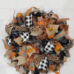 May include: A decorative fall wreath featuring a mix of ribbons in black and white buffalo check, leopard print, and burlap. Orange ribbons with pumpkin designs are interspersed throughout the wreath, creating a festive autumn theme.