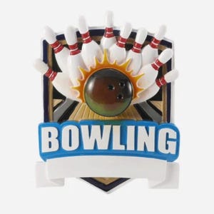 May include: A decorative bowling ornament featuring a dark brown bowling ball striking white pins with red stripes. A yellow sunburst graphic sits behind the ball. The word BOWLING is printed in large white letters on a blue banner above a blank white space.