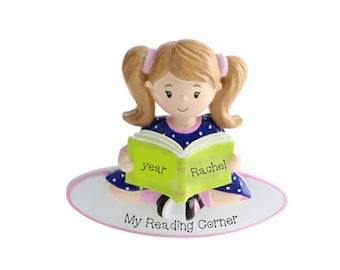 May include: A figurine of a girl with blonde pigtails wearing a blue dress with white polka dots, reading a green book. The book reads "Year Rachel". The base reads "My Reading Corner".