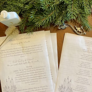 May include: A booklet titled 'Advent Wreath Prayers' with text and illustrations, a bottle, and a decorative metal piece. The booklet contains prayers for the blessing of the wreath and the first Sunday of Advent. Green pine branches form the background.