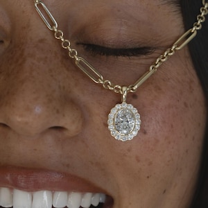 May include: A gold paperclip chain necklace featuring an oval-cut center stone surrounded by a halo of small round white stones. The pendant is suspended from the elongated rectangular links of the gold chain, creating a classic jewelry piece.