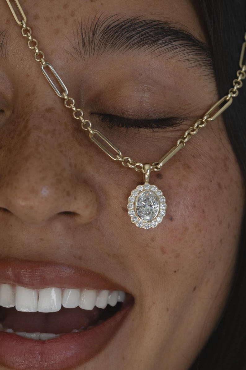 May include: A gold paperclip chain necklace featuring an oval-cut center stone surrounded by a halo of small round white stones. The pendant is suspended from the elongated rectangular links of the gold chain, creating a classic jewelry piece.