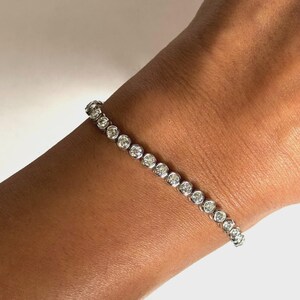 May include: A silver-toned tennis bracelet featuring a continuous row of round, clear, faceted stones set in individual circular metal bezels. The bracelet is designed with a flexible link construction that wraps around the wrist in a straight line.