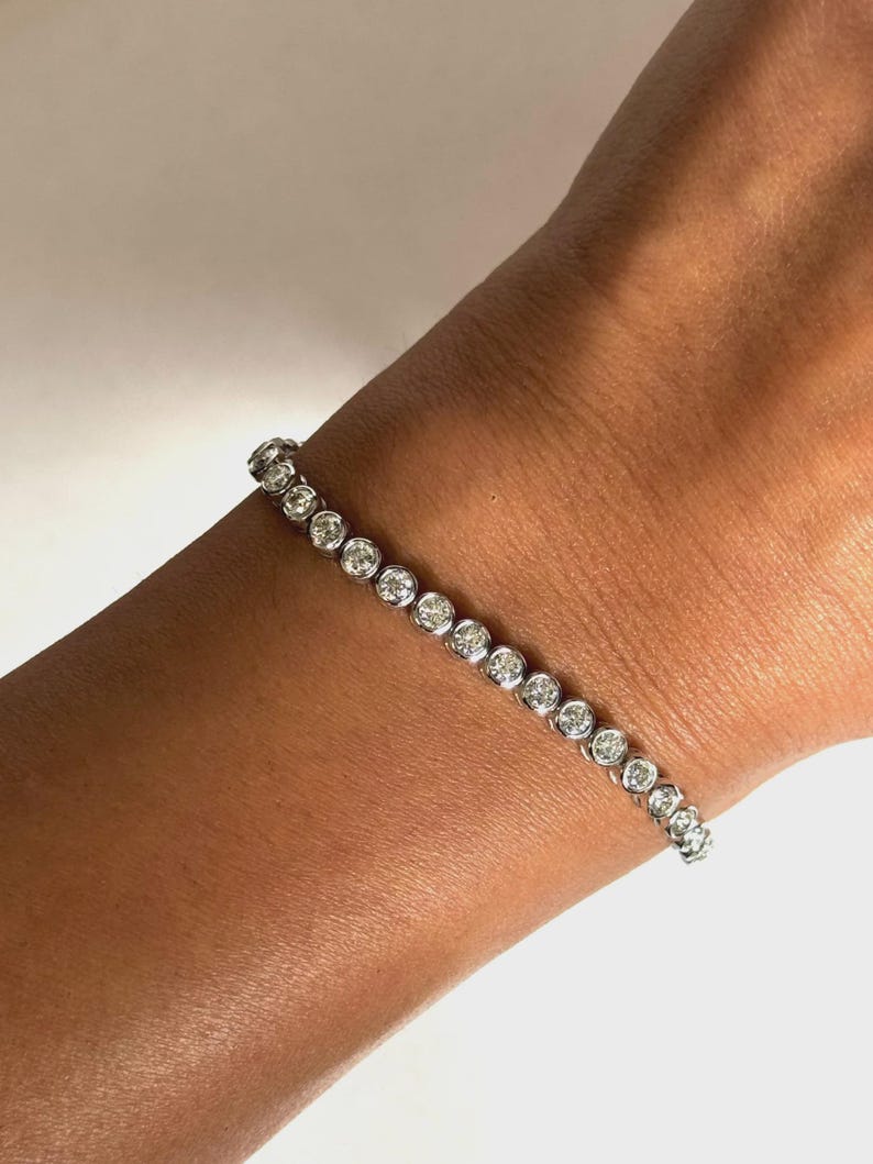 May include: A silver-toned tennis bracelet featuring a continuous row of round, clear, faceted stones set in individual circular metal bezels. The bracelet is designed with a flexible link construction that wraps around the wrist in a straight line.