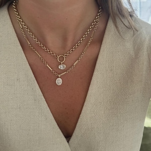 May include: A layered gold necklace set featuring a shorter chain with a round link and a single oval pendant, and a longer chain with elongated bar links and a smaller oval pendant encrusted with small clear stones, worn against a beige top.