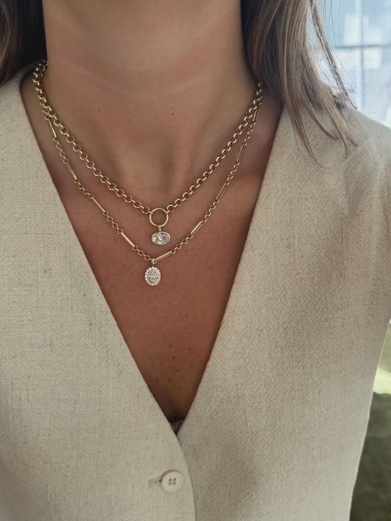 May include: A layered gold necklace set featuring a shorter chain with a round link and a single oval pendant, and a longer chain with elongated bar links and a smaller oval pendant encrusted with small clear stones, worn against a beige top.