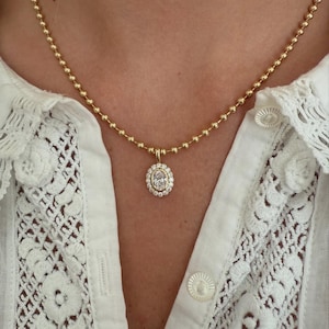 May include: A gold ball chain necklace featuring an oval-shaped pendant. The pendant has a central oval stone surrounded by a halo of smaller round stones. The necklace is worn against a white shirt with lace detailing and round buttons.