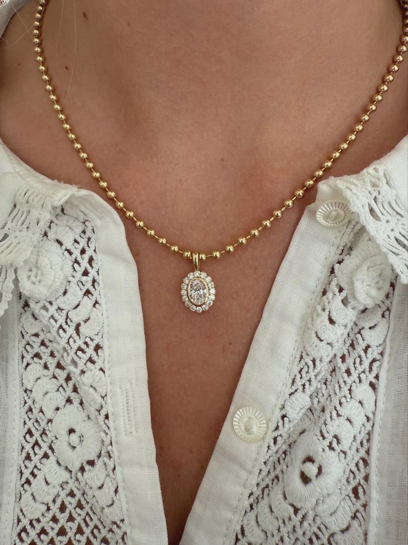 May include: A gold ball chain necklace featuring an oval-shaped pendant. The pendant has a central oval stone surrounded by a halo of smaller round stones. The necklace is worn against a white shirt with lace detailing and round buttons.
