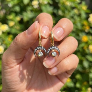 May include: A pair of gold hoop earrings featuring a crescent moon design. The black crescent is encrusted with small, sparkling stones, and a larger, round, clear stone hangs from the center. The earrings are held against a blurred green and yellow background.