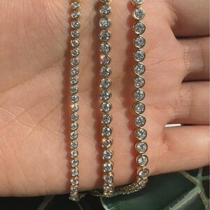 May include: Three gold-toned tennis bracelets featuring a continuous row of round, clear, bezel-set stones. Each bracelet displays a uniform design with metallic gold settings surrounding the circular, faceted, reflective white stones.