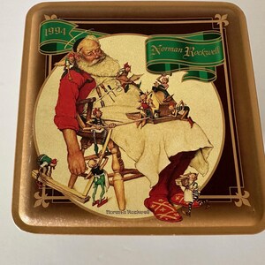 Könnte beinhalten: Rechteckige, goldfarbene Metallschale mit einer Norman Rockwell-Illustration von Santa Claus, umgeben von Elfen. Die Illustration befindet sich vor einem cremefarbenen Hintergrund, mit dem Text "1994" und "Norman Rockwell" auf der Schale.