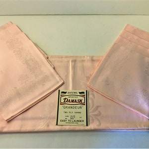 Puede incluir: Un juego de mantelería de damasco rosa con un patrón floral. La mantelería está doblada y dispuesta sobre una superficie blanca. La etiqueta dice "Bardwil Imported Damask 'Grandeur' Two Ply Yarns Easy to Launder".