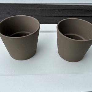 May include: Two brown ceramic plant pots on a white surface. The pots are cylindrical and have a smooth finish.